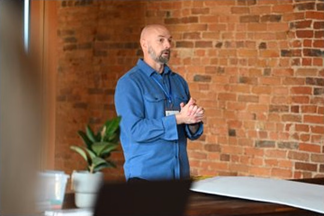 Niels in a blue shirt speaking in front of an exposed brick wall, hands clasped, mid-thought