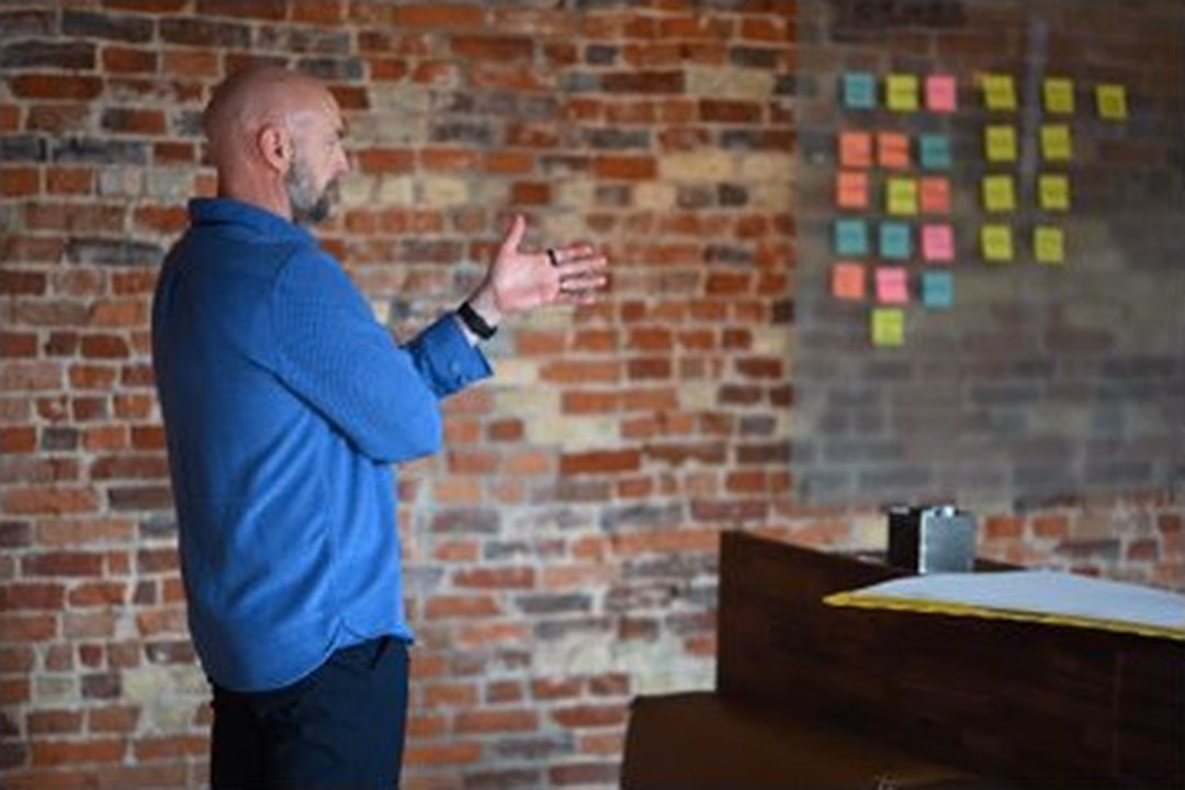 Niels mid-sentence in a blue shirt, hands open, in front of an exposed brick wall