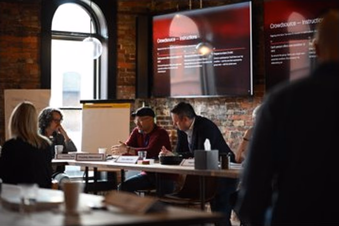 A leadership team at long wooden tables in a brick-walled room, seen from the back, Niels standing at the front