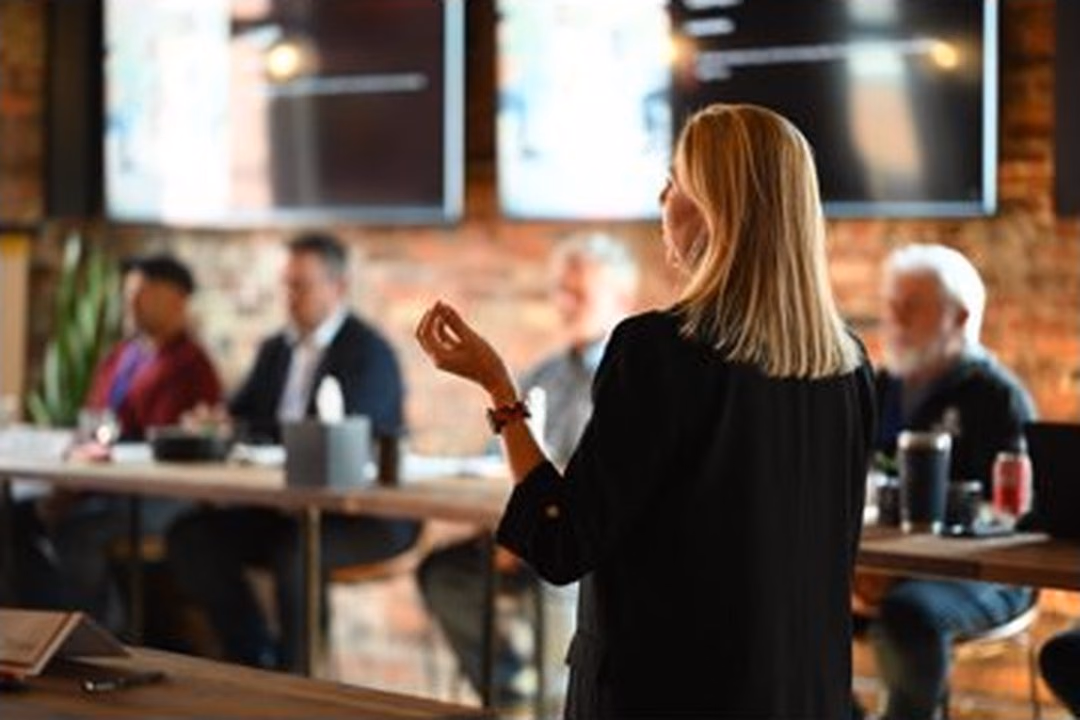 Fleur presenting to a room, gesturing, participants at tables in the foreground