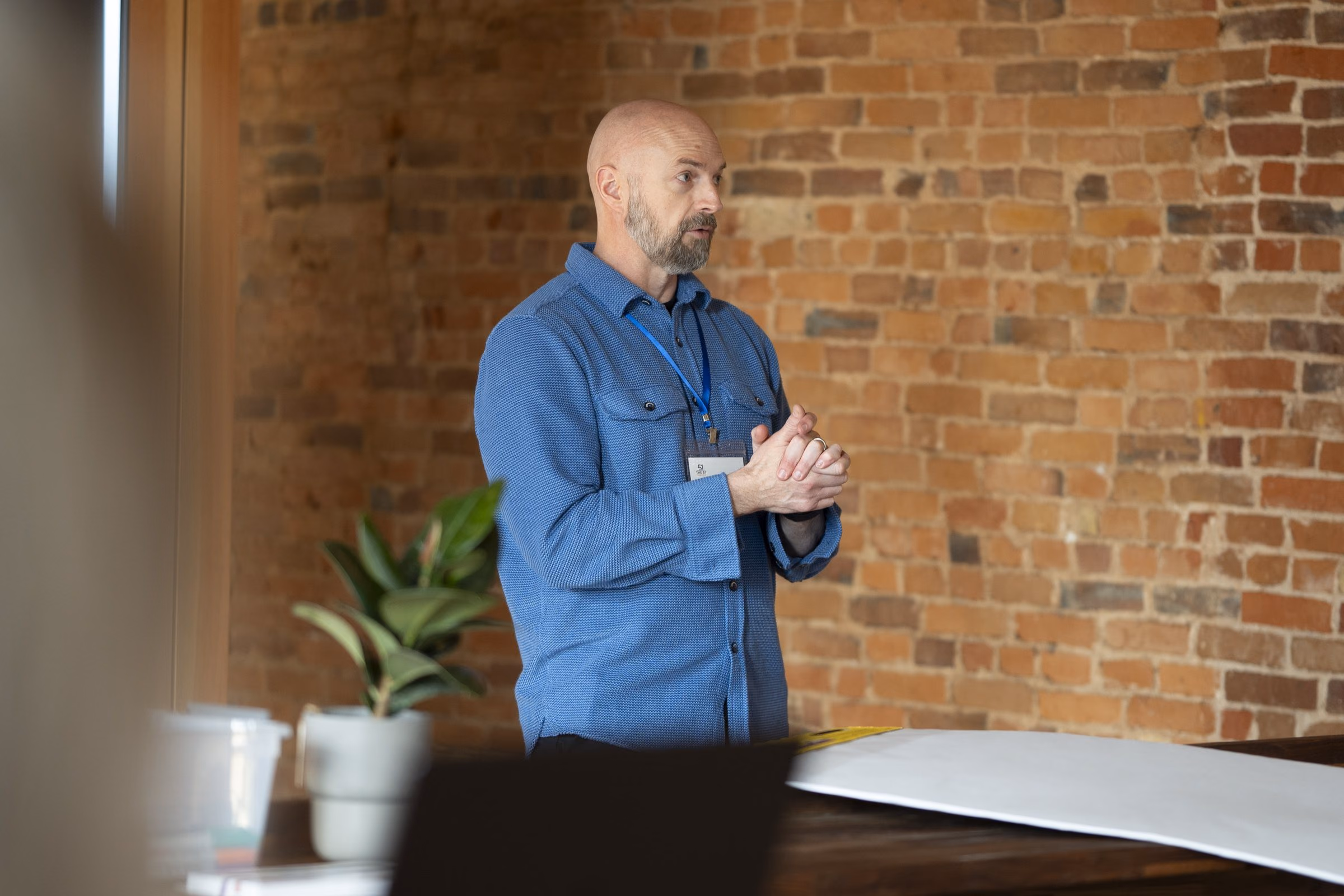 Niels in a blue shirt at the front of a brick-walled room, mid-thought, a plant beside him