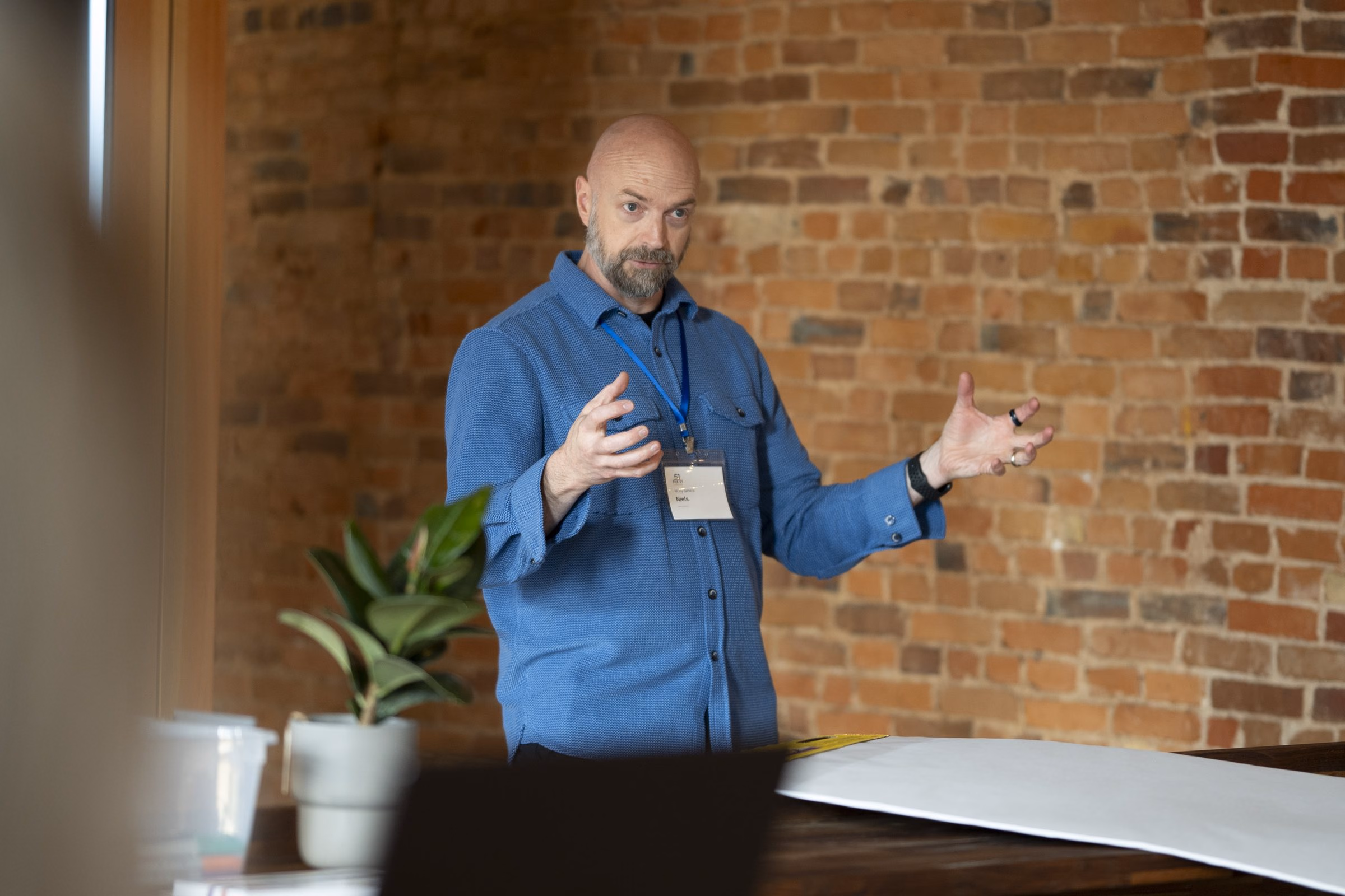 Niels mid-sentence in a blue shirt, both hands open, exposed brick behind him