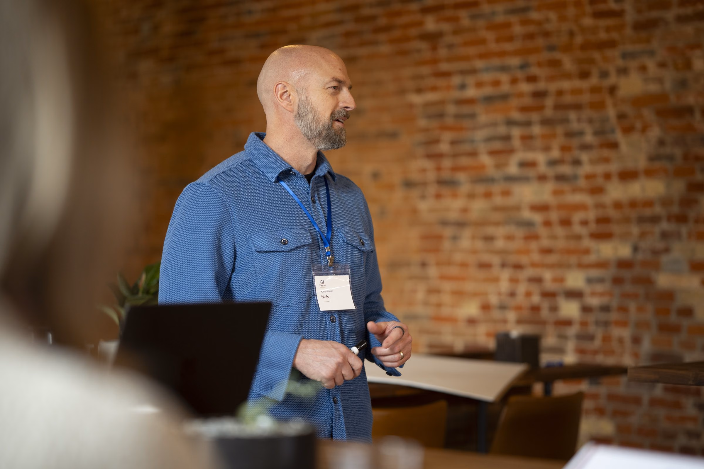 Niels at a laptop at the front of a brick-walled room, hands open, mid-sentence