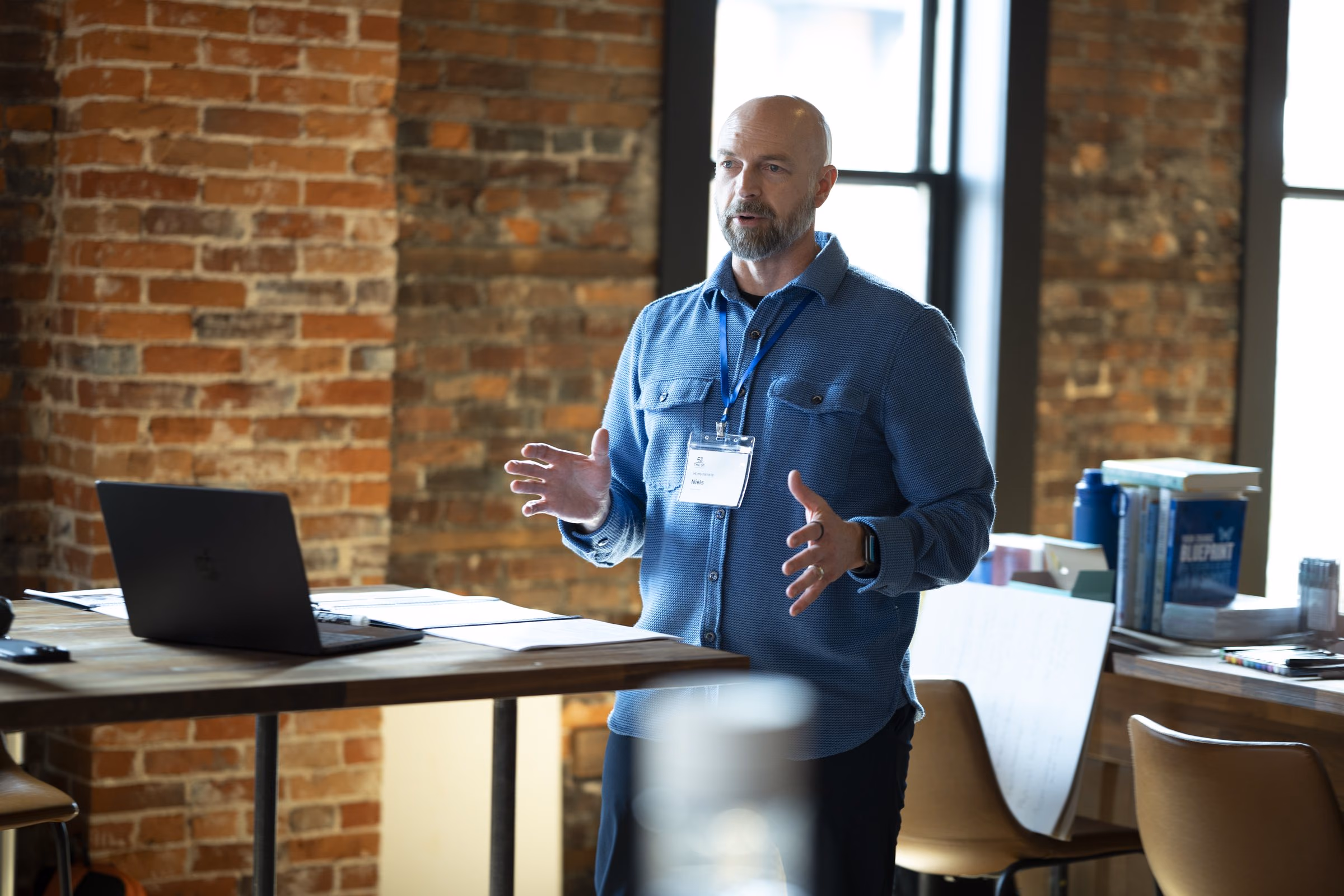 Niels standing at a laptop at the front of a brick-walled room, gesturing to the team