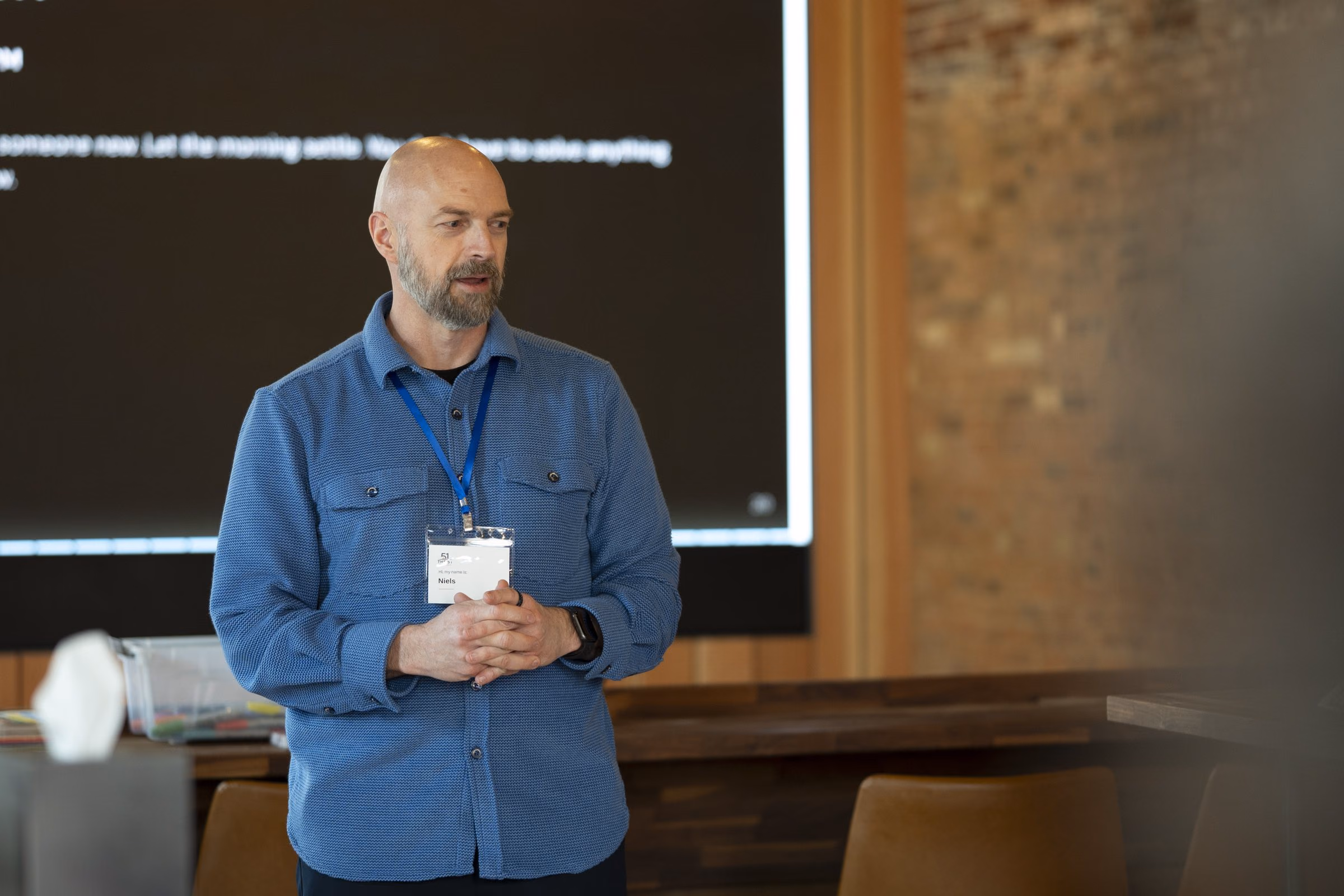Niels standing in a brick-walled room with a coffee in hand, relaxed, looking out at the room