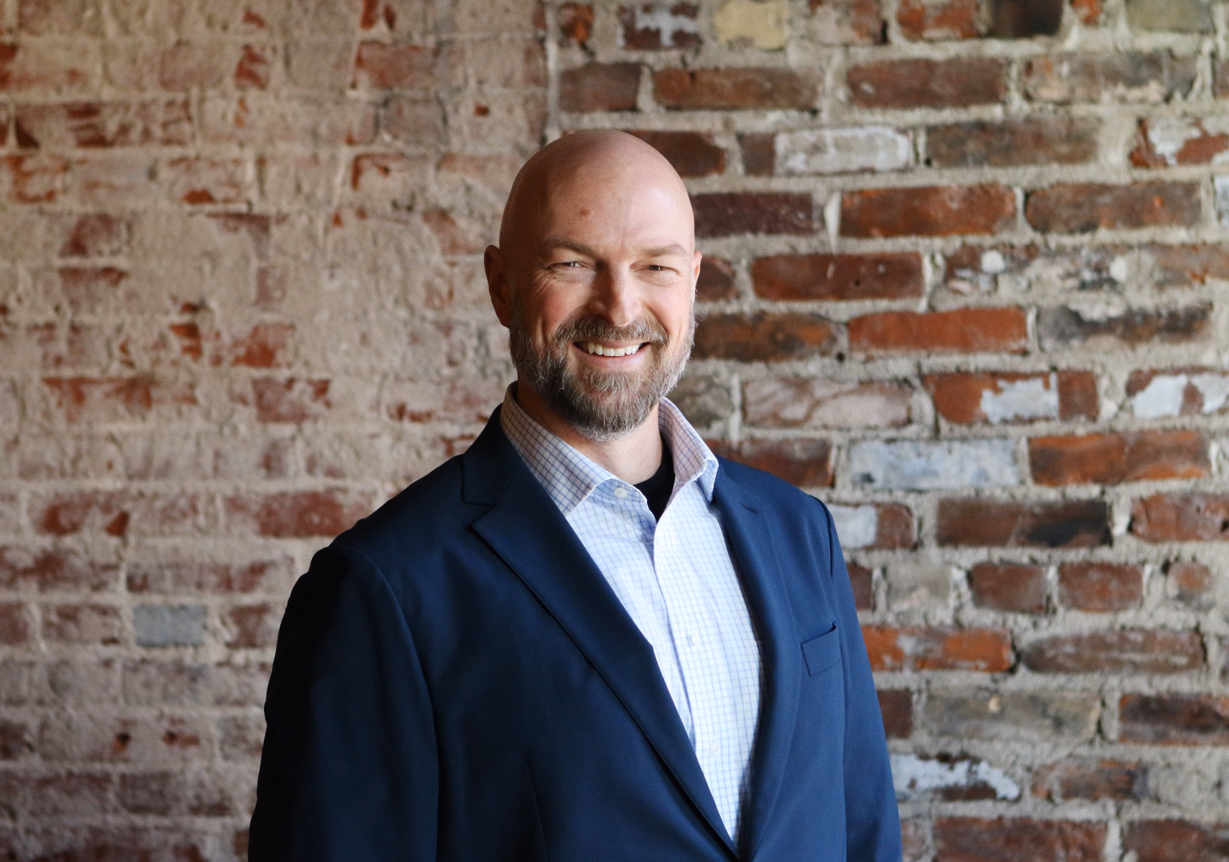 Niels in a navy blazer, three-quarter to the camera, relaxed smile, brick wall behind