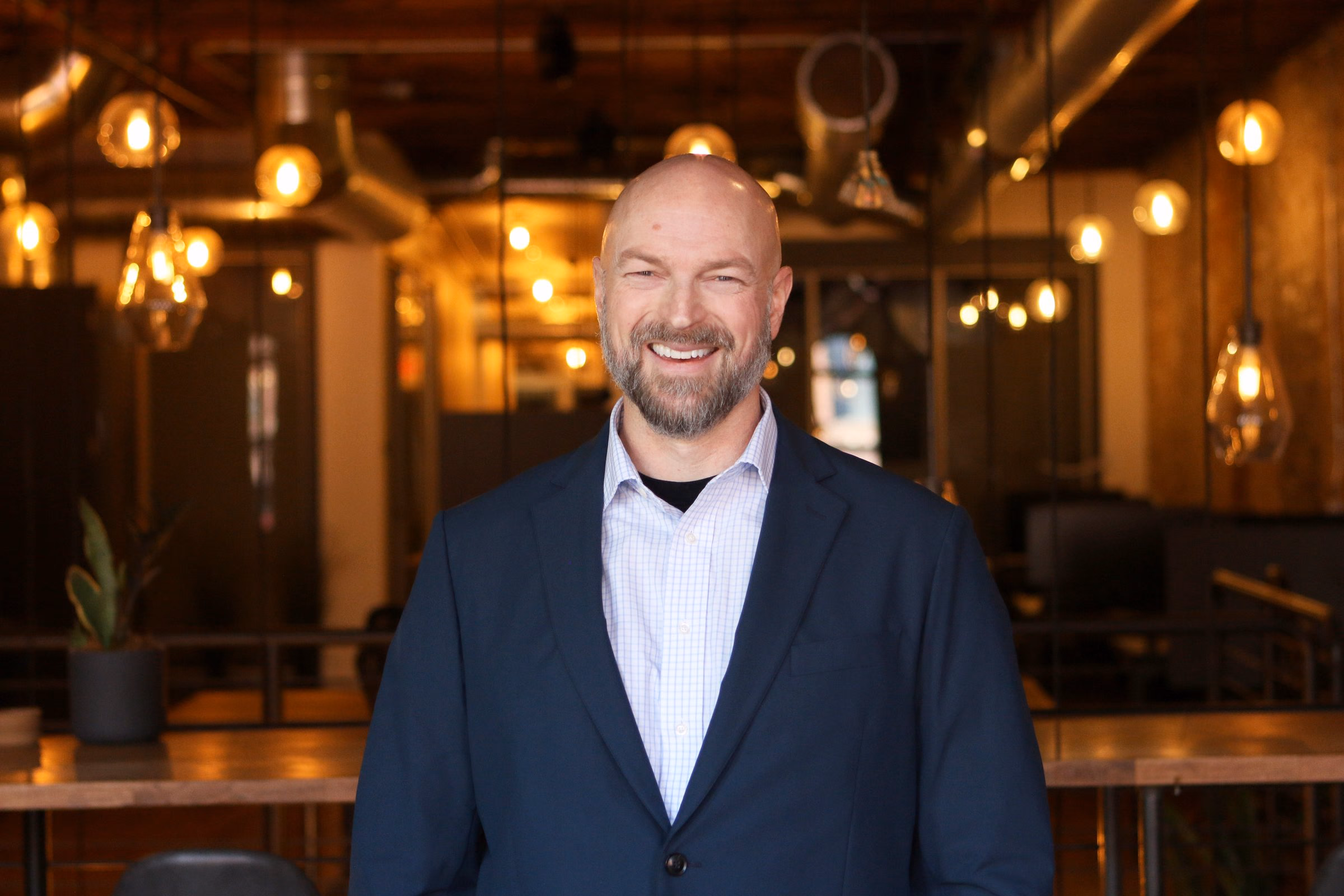 Niels standing in a navy blazer, smiling at the camera, warm lights behind him