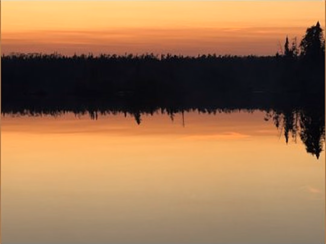 A still lake at dusk in the Boundary Waters, pines on the far shore, the sky fading from gold to navy